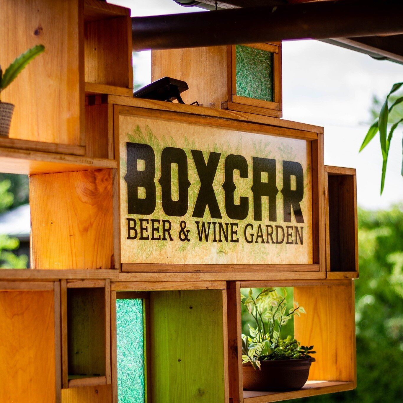 Wooden sign reading “BOXCAR Beer & Wine Garden” among rustic shelves and greenery