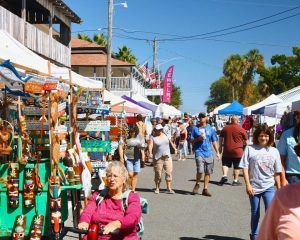 Crowded outdoor street market with white tents, shoppers, and colorful hanging crafts on a sunny day