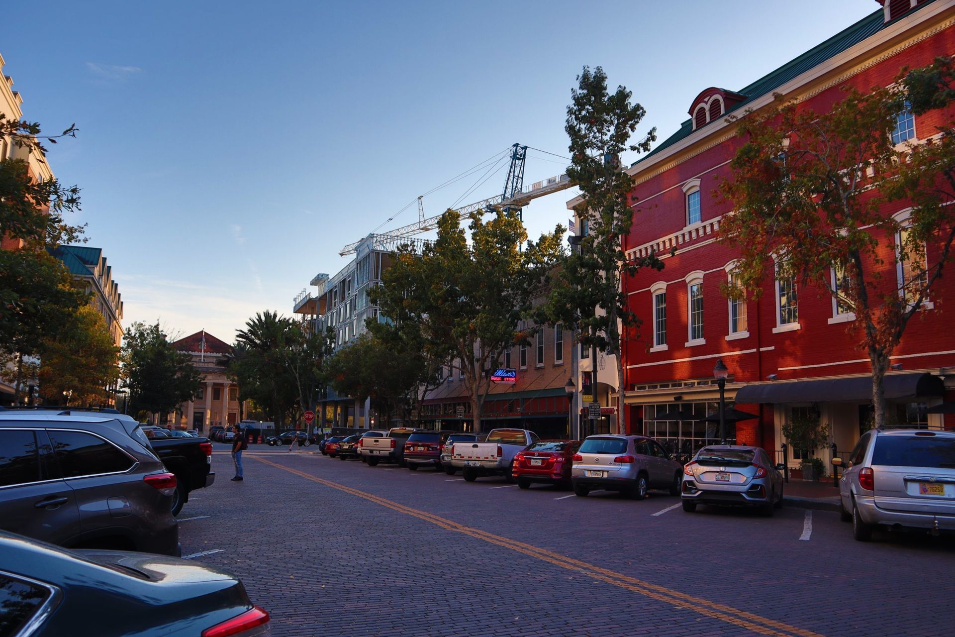 Quiet cobblestone street with parked cars, red brick buildings, and trees at dusk