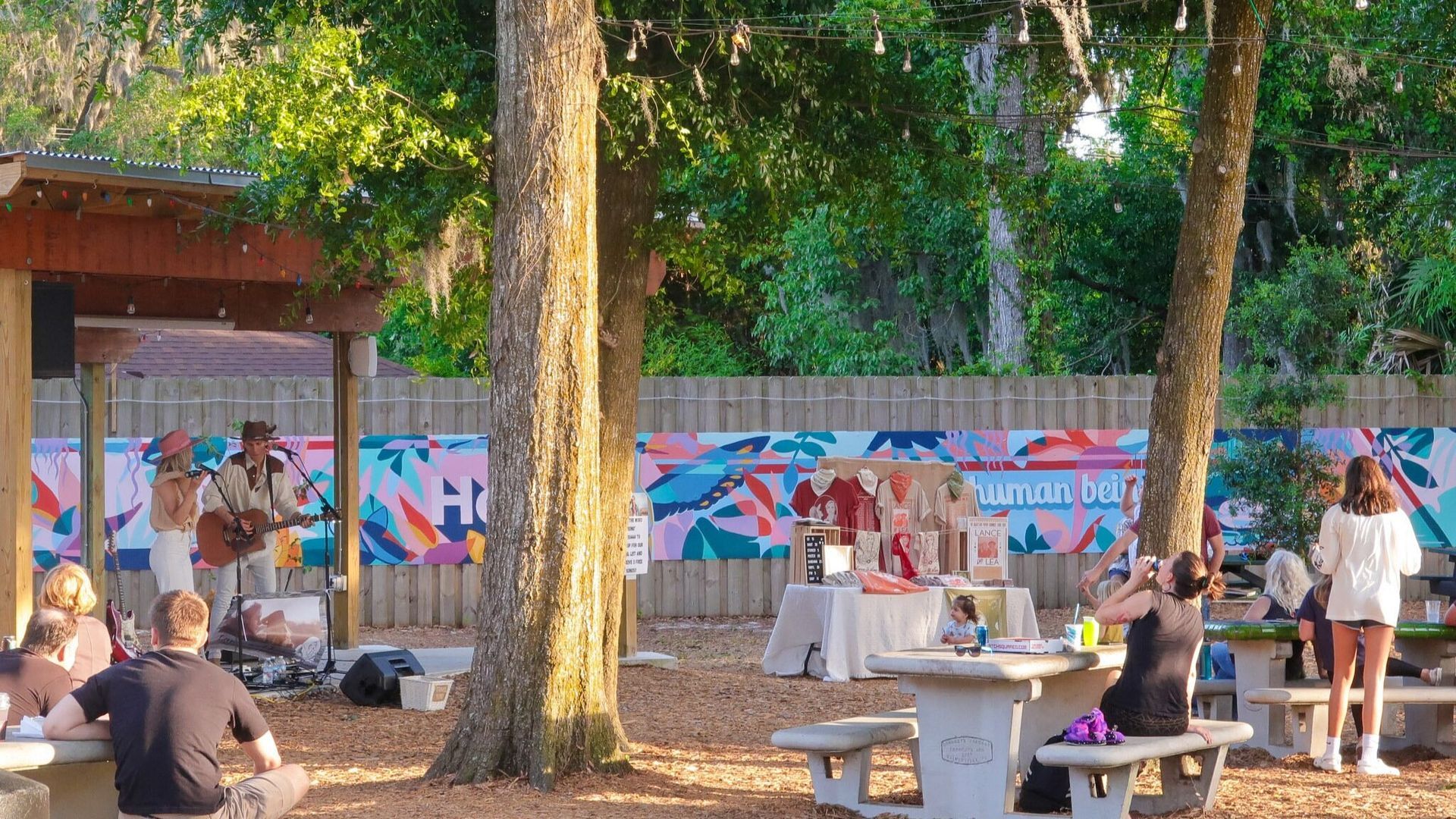 Outdoor park gathering with people at tables under trees beside a colorful mural wall.