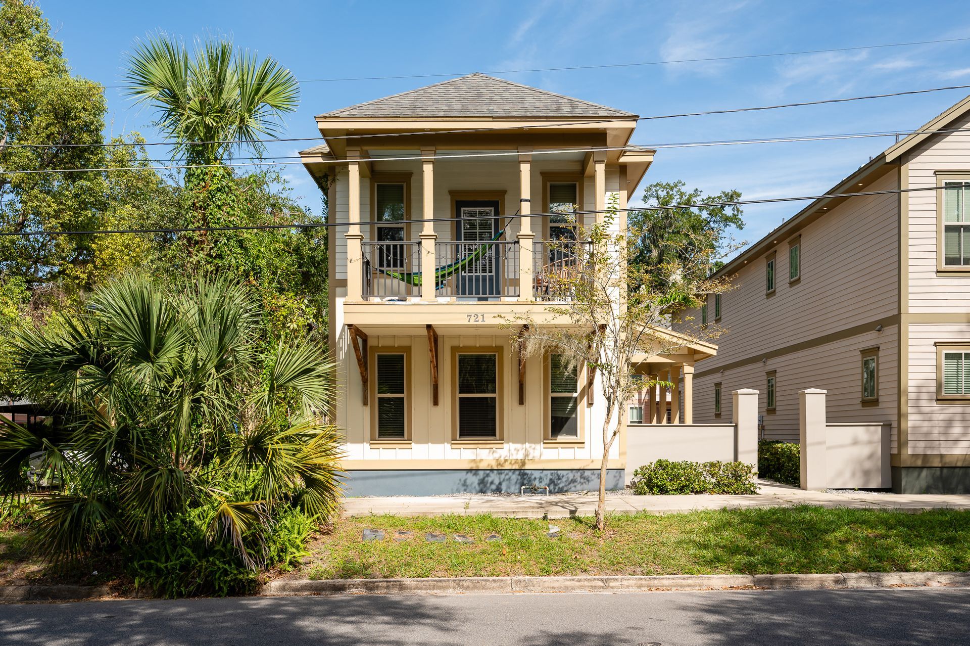 Two-story beige house with a front porch, columns, and balconies on a sunny street with trees nearby