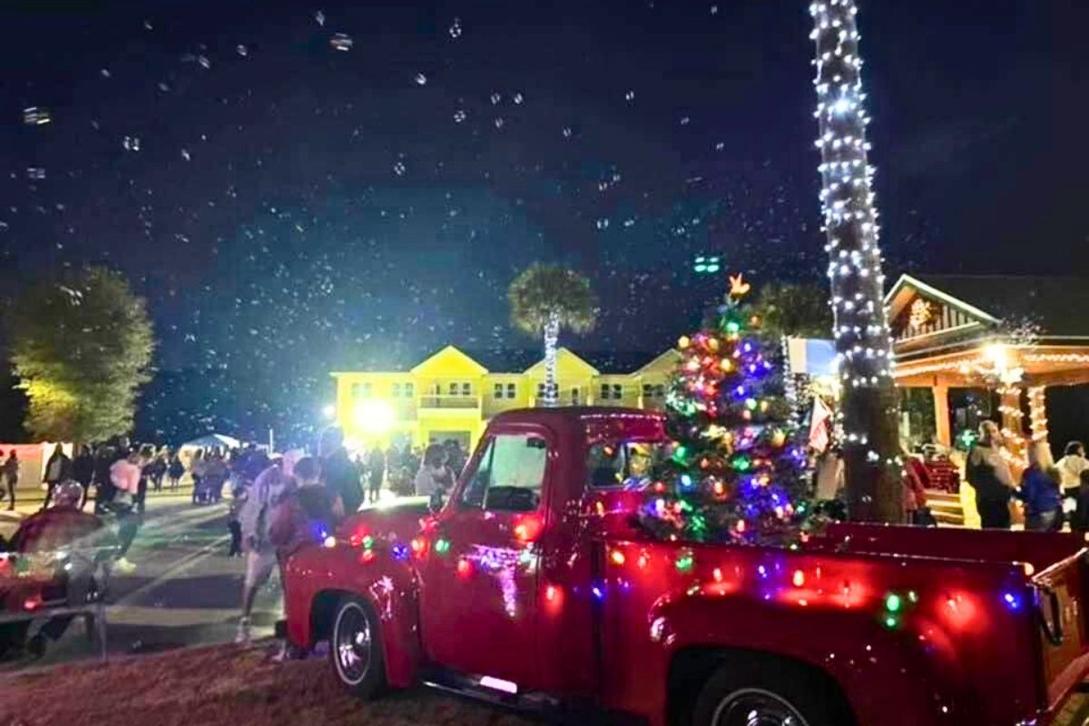 Red pickup truck decorated with holiday lights at a nighttime Christmas parade, with a lit tree nearby