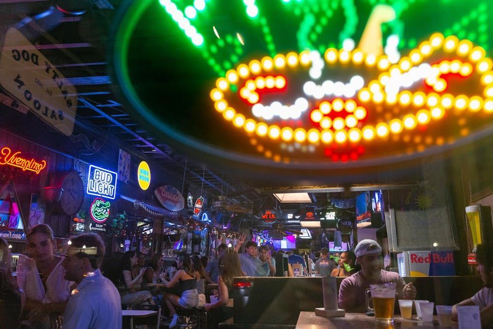 Neon-lit bar interior with people seated at tables and colorful signs overhead