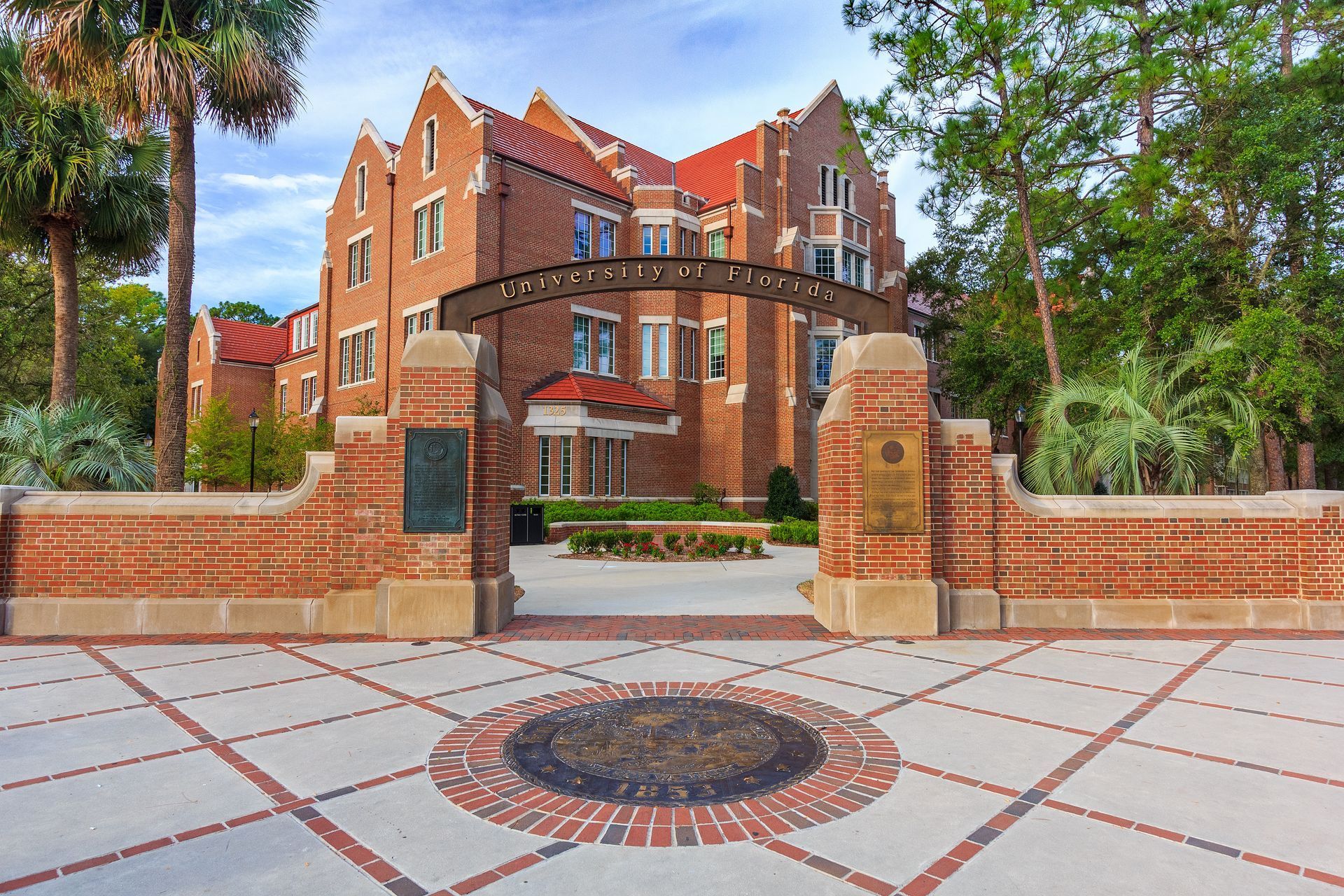 Brick mansion behind a gated entrance with red tile accents and trees lining the street