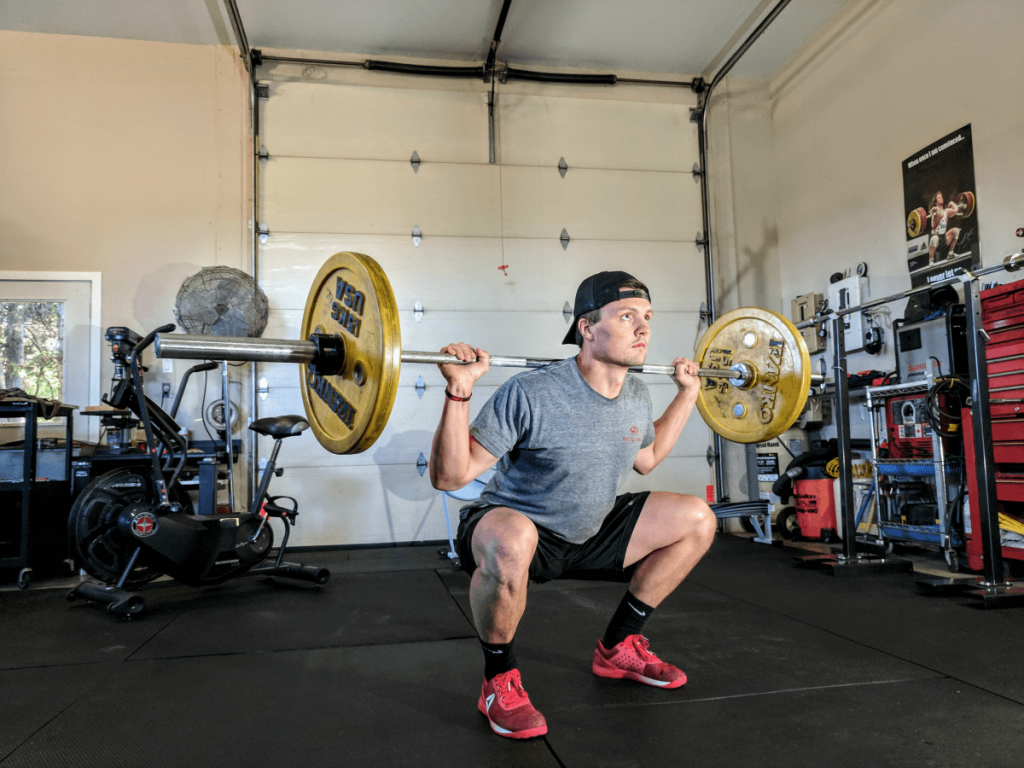 A man is squatting with a barbell in a garage.