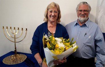 Smiling couple beside a gold menorah, holding a bouquet in a simple indoor setting