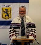 Man standing at a podium in front of an Israeli flag, wearing a striped tallit and tie.