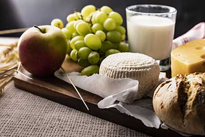 Apple, green grapes, cheese, milk, and bread on a wooden table with a knife and wrapped cheese wedge