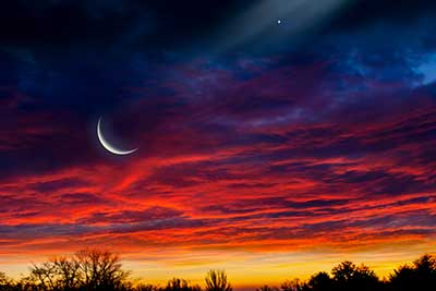 Crescent moon over a vivid red-purple sunset sky with dark tree silhouettes on the horizon