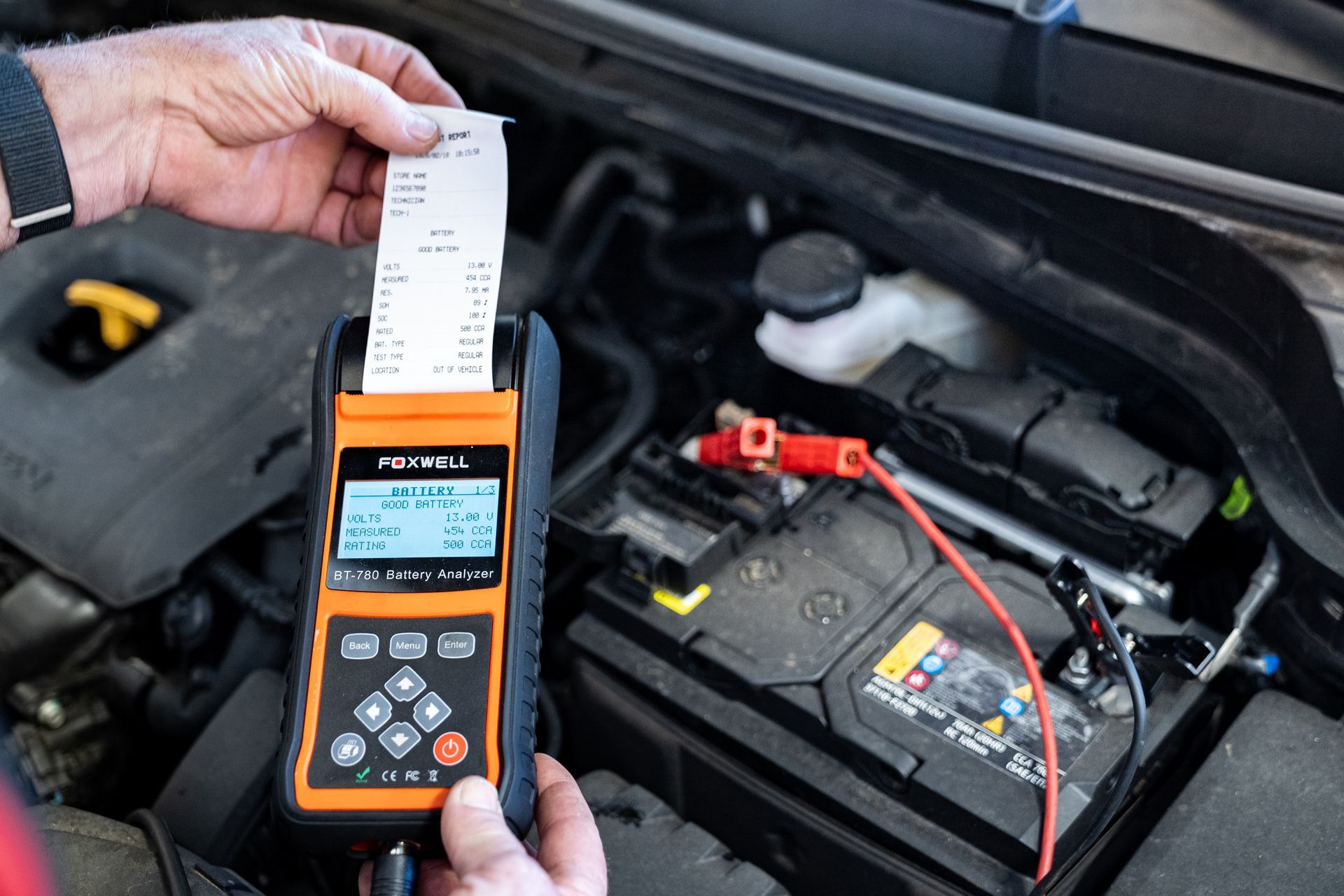 Handheld battery tester printing a receipt while connected to a car battery under the hood