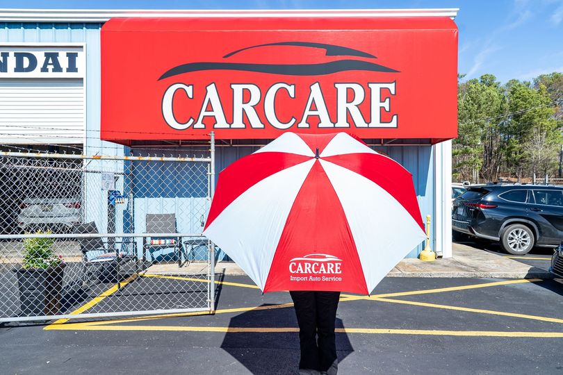 Person holding a red-and-white CARCARE umbrella in a parking lot outside a car care shop