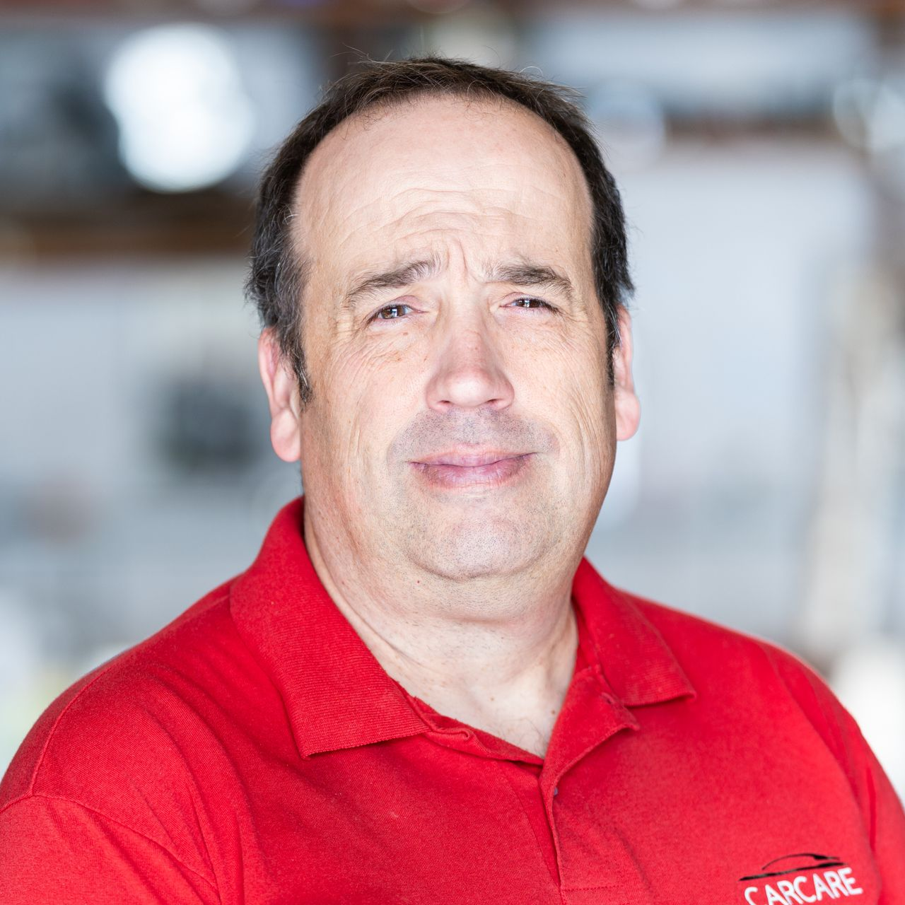 Man in a red polo shirt with a neutral expression, standing against a blurred background