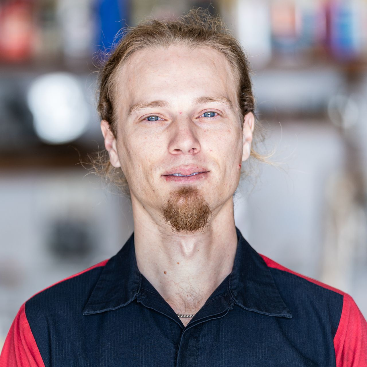 Portrait of a person in a dark navy shirt with red shoulders, standing indoors against a blurred background