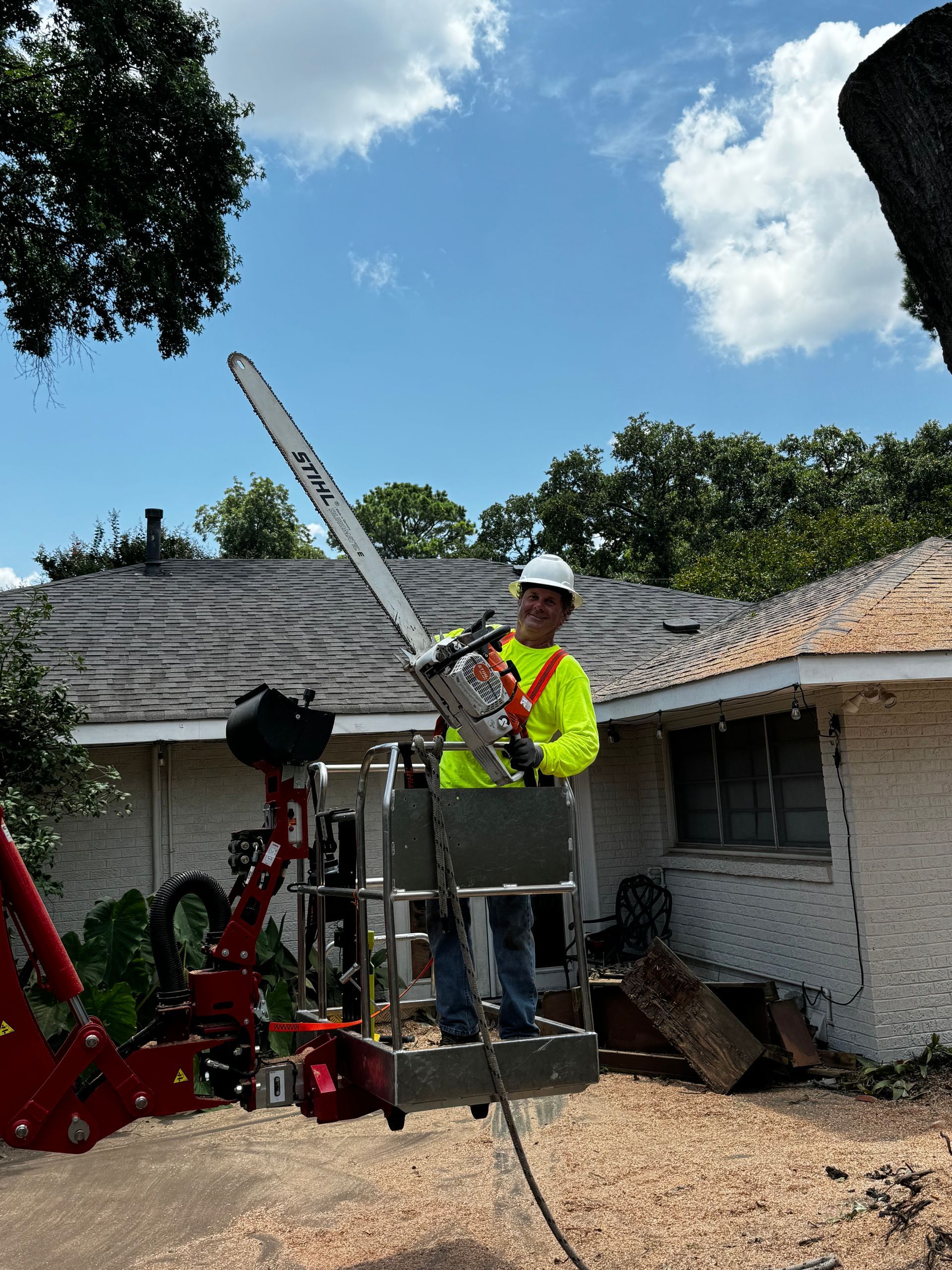 Arborist in lift using a chainsaw, trimming a tree near a house, sunny day.