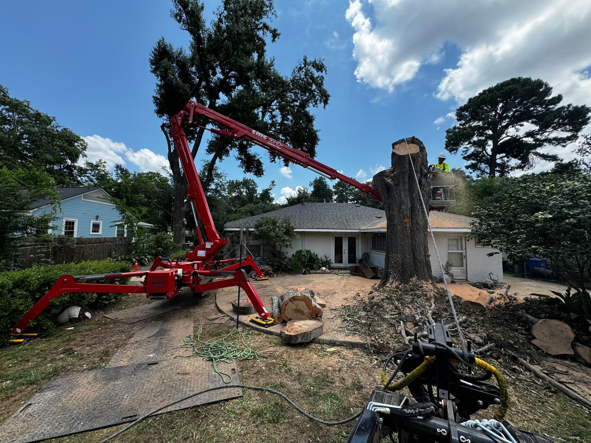 Red crane removing a large tree near a house. Branches and wood debris scattered on the ground under a blue sky.