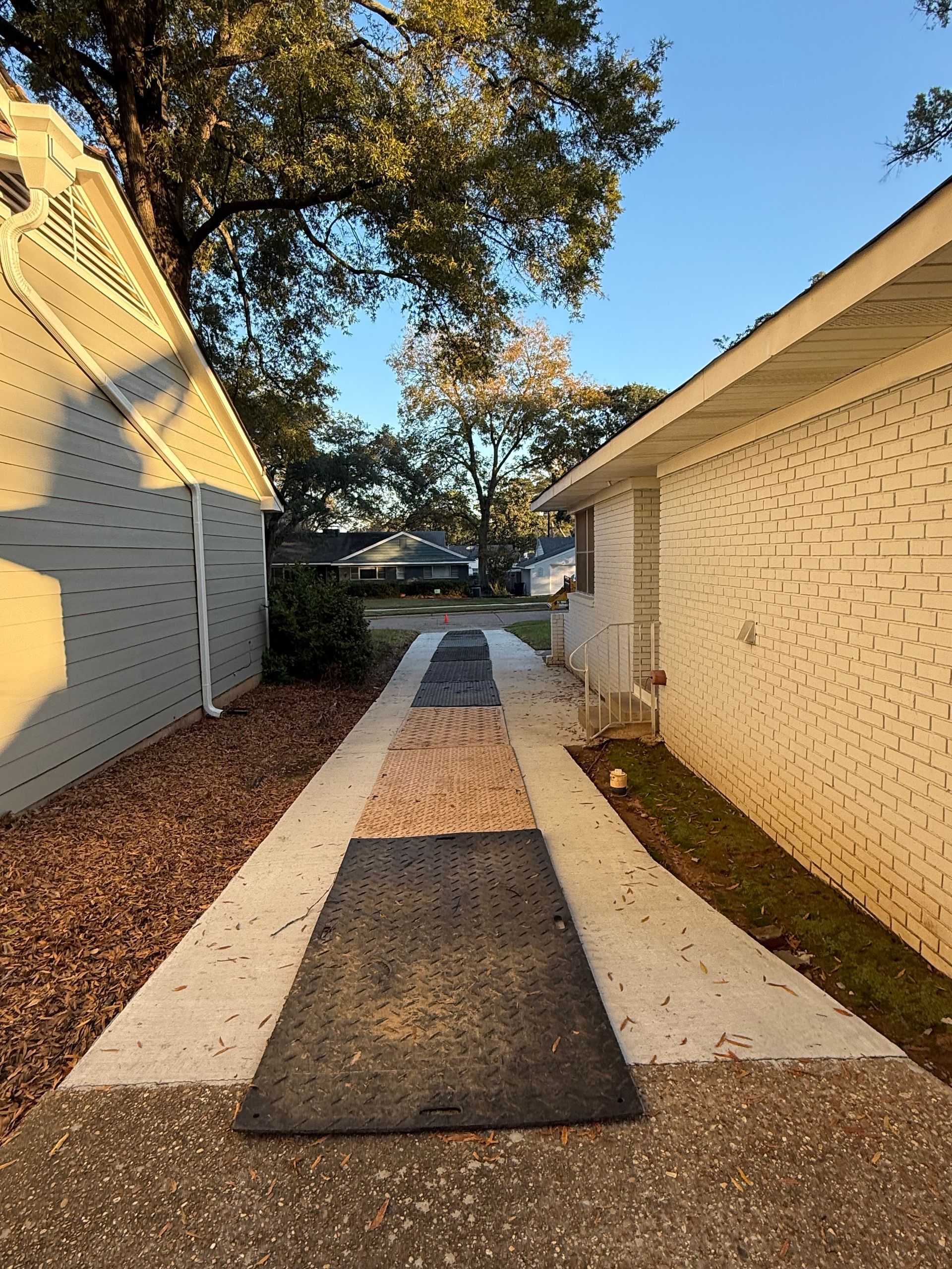 Concrete pathway with rubber mats between two buildings, trees in the background under a blue sky.
