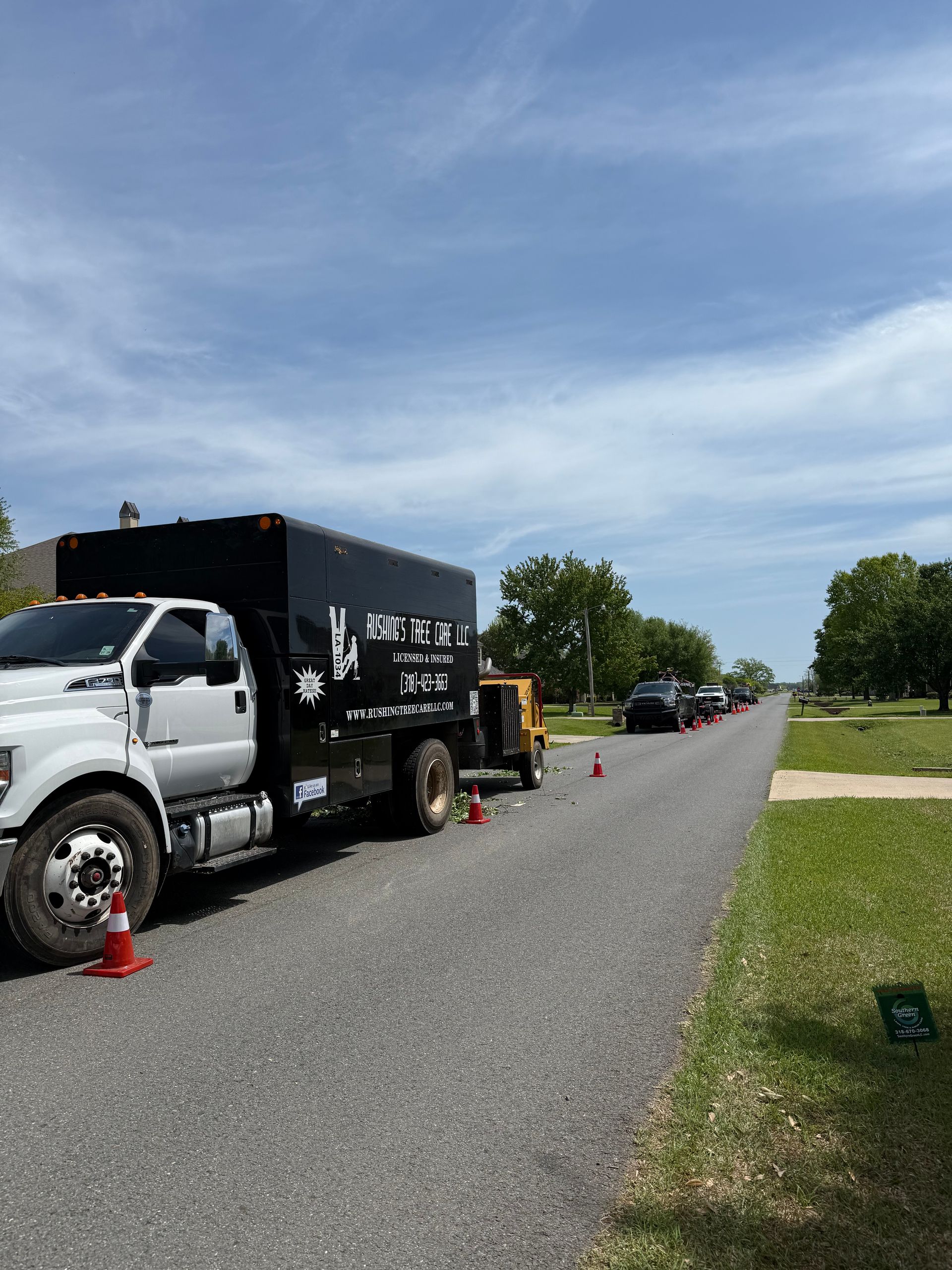 White truck with a black container parked on a road lined with orange cones, other vehicles further down the road, clear sky.