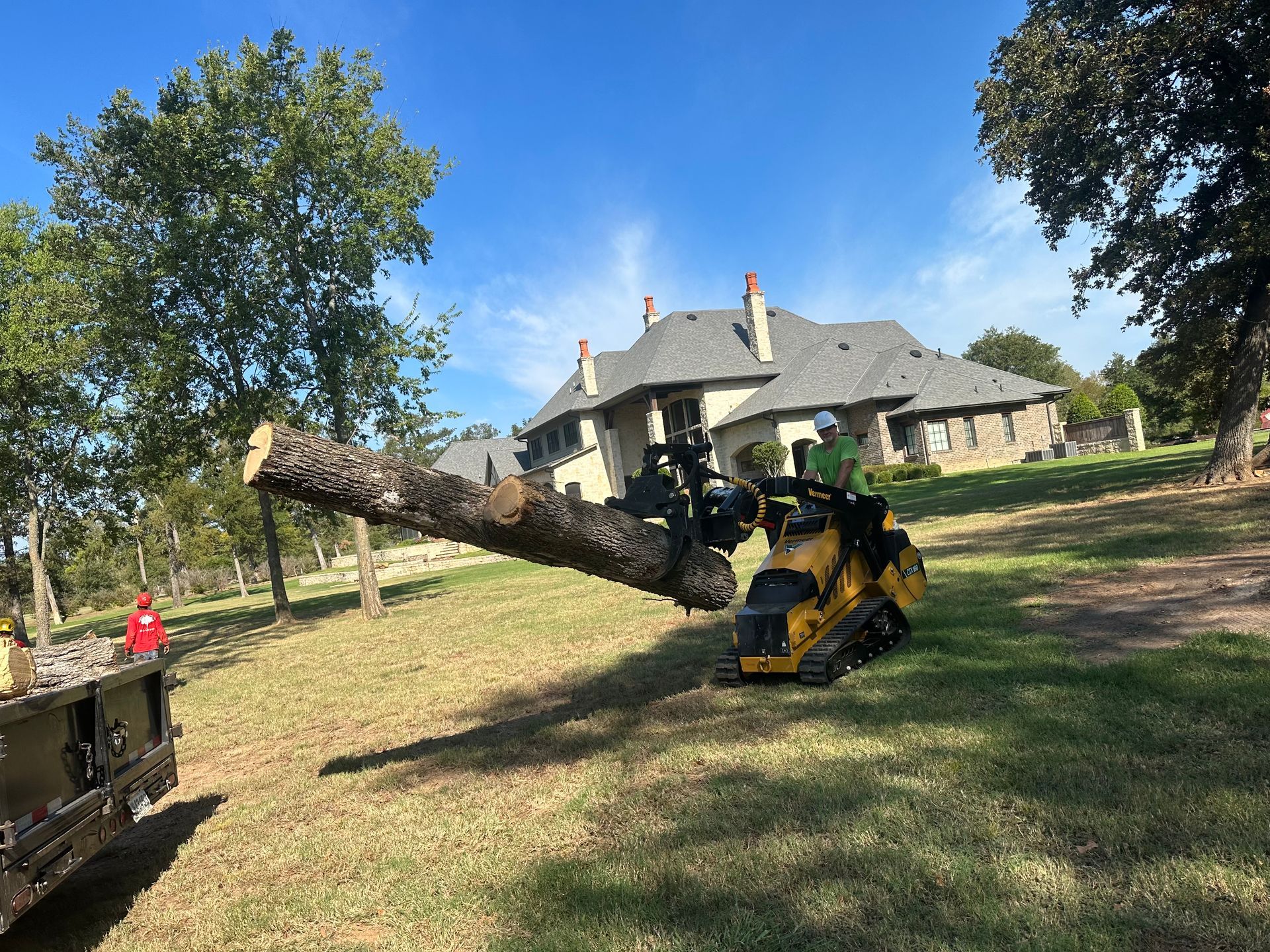 Mini excavator holding tree trunk, placing it near a wood chipper on a grassy hillside, house in background.