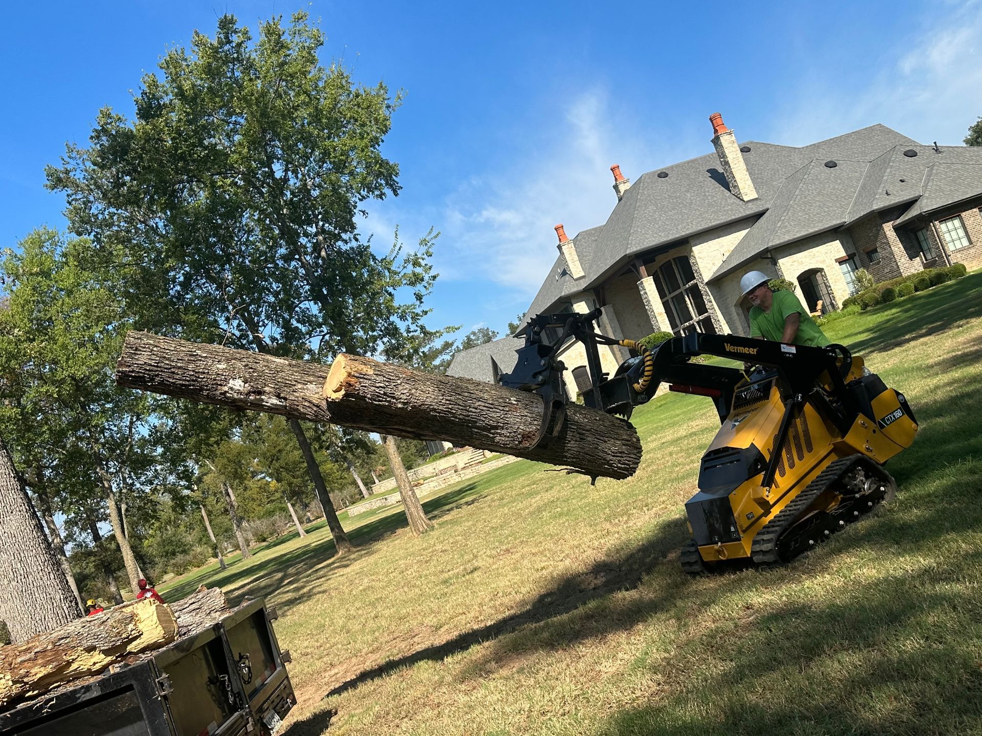 Yellow mini-excavator with a log, operator in a yard, house in background.