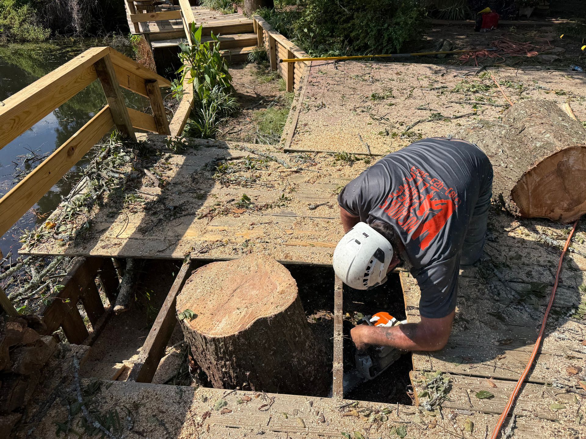 Man cutting a tree stump on a wooden deck, near a pond with a chainsaw.