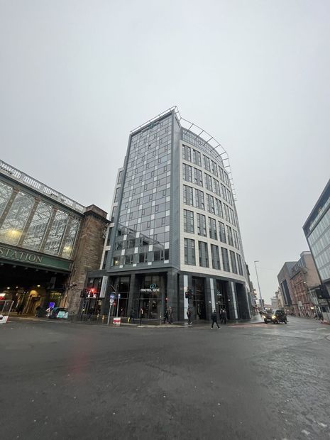 Modern high-rise building on a street corner, next to a vintage train station. Overcast sky.