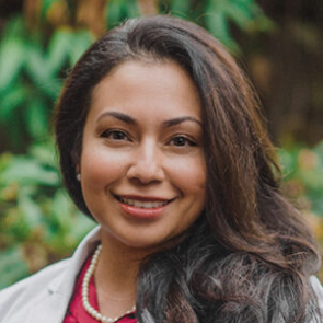 A smiling person with long, dark hair, wearing a white blazer over a reddish top and a pearl necklace, outdoors.