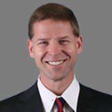 Headshot of a smiling person wearing a dark suit, white collared shirt, and a red patterned tie against a grey background.