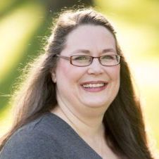 A smiling person with long dark hair and glasses wears a gray shirt, set against a soft, sunlit green background.