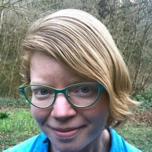 A person with blonde hair and teal-rimmed glasses smiles slightly in a forest setting, wearing a blue shirt.