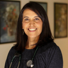 A smiling professional wearing a stethoscope over a dark shirt, standing in front of two framed paintings.