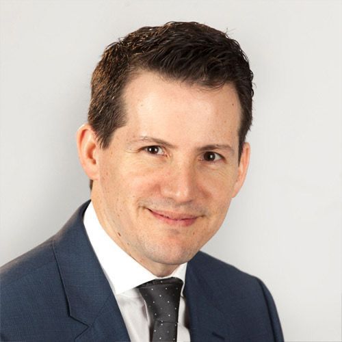 Head-and-shoulders portrait of a person wearing a navy blue suit, white shirt, and dark patterned tie against a light wall.