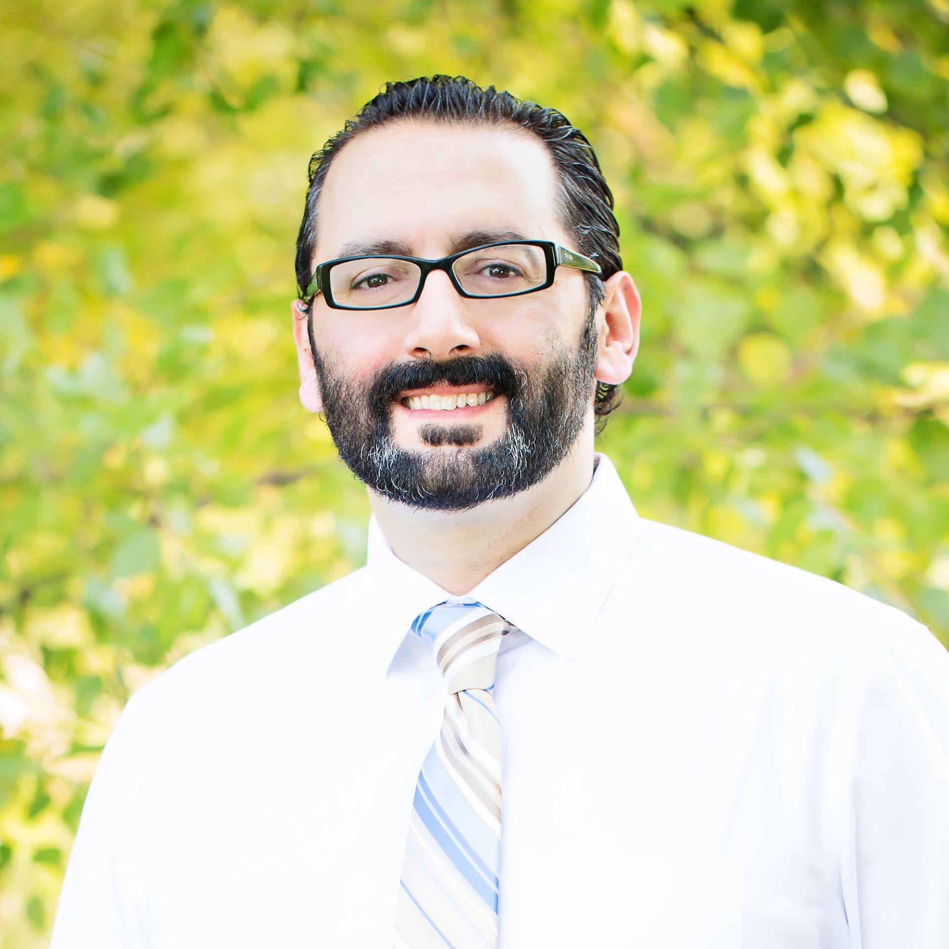 A smiling person with glasses and a beard wearing a white dress shirt and striped tie in front of a blurry green forest.