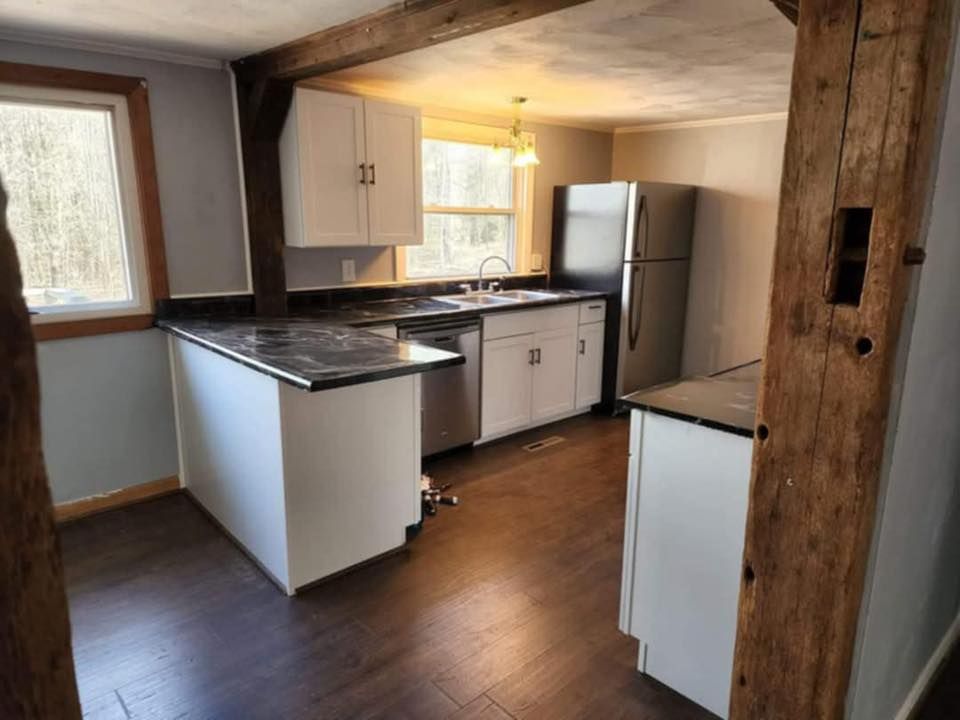 Small rustic kitchen with white cabinets, dark countertops, wood beams, and a window-lit dining area