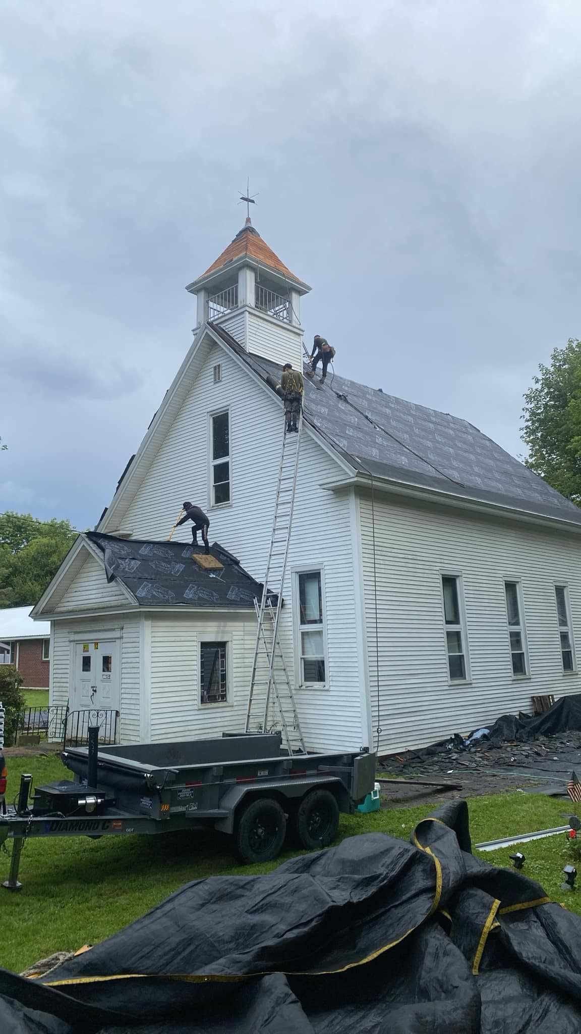 Damaged white church with a small steeple, tarp-covered debris in the foreground, and stormy sky