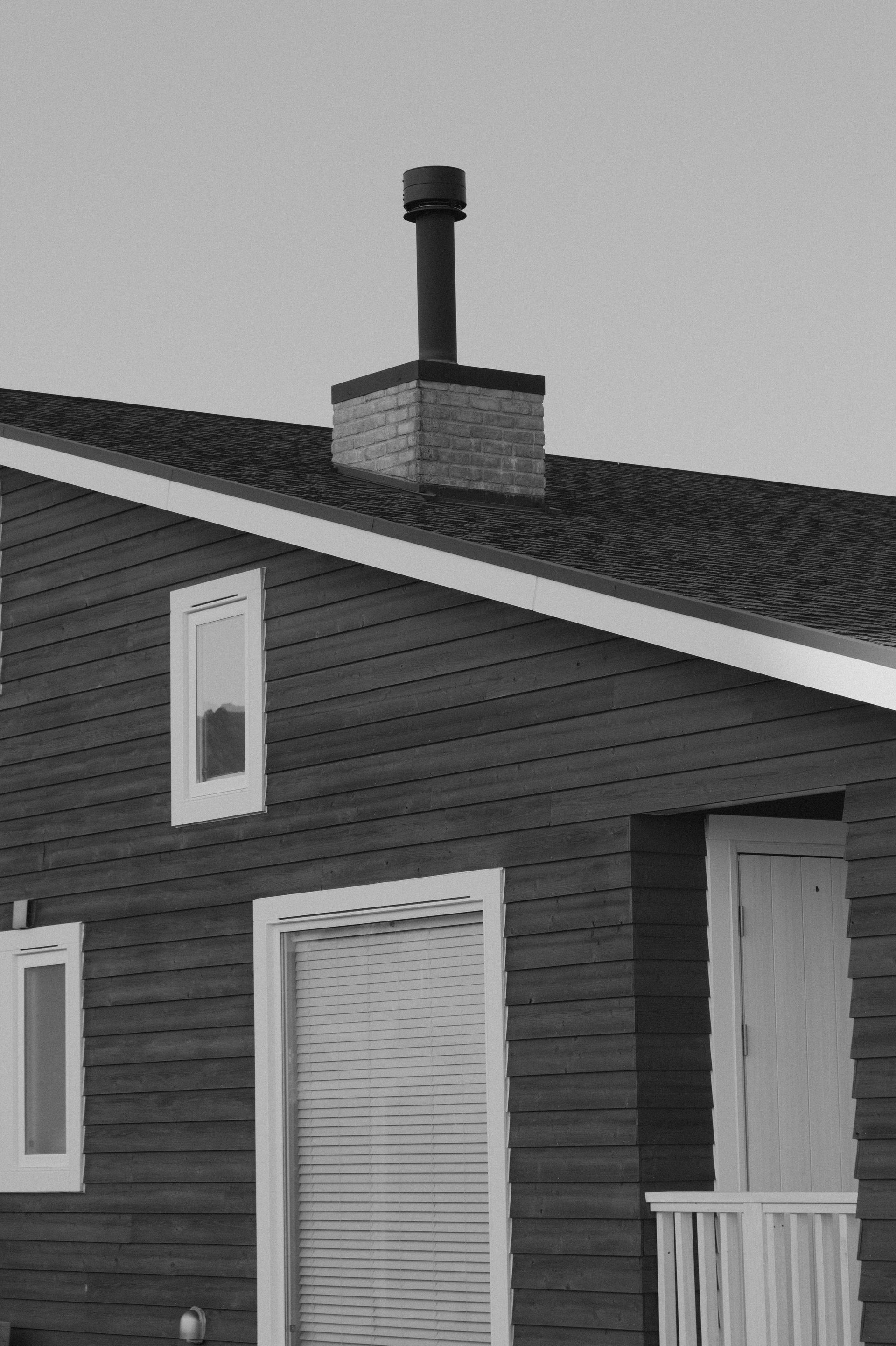 Side of a house with dark siding, white windows and door, and a rooftop chimney against a gray sky