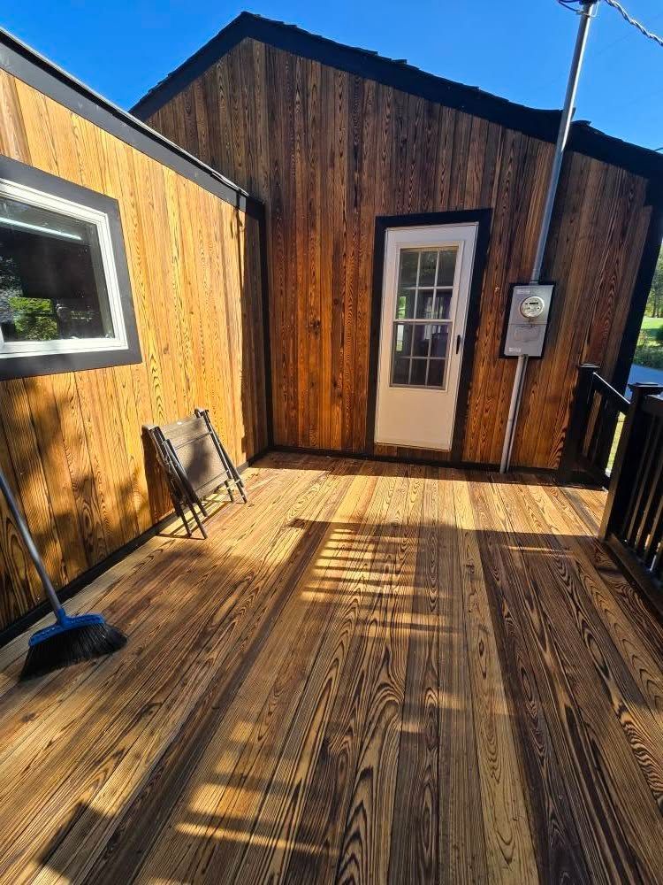Wooden deck with a small dark cabin wall, white door, window, and strong sunlight casting shadows