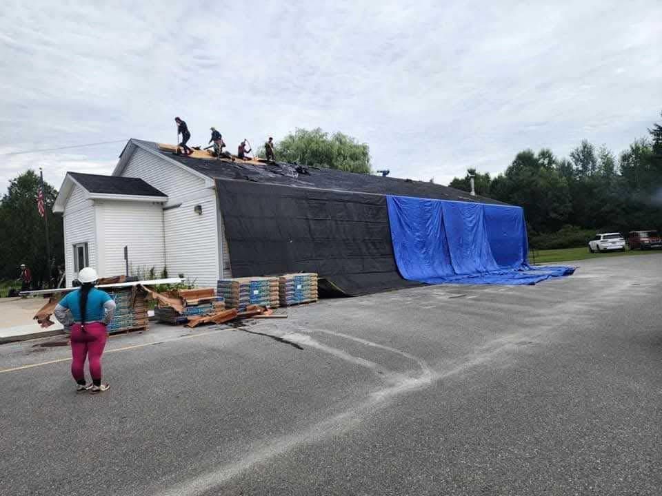 Workers on a roof with tarps covering part of the building and pallets stacked below outdoors