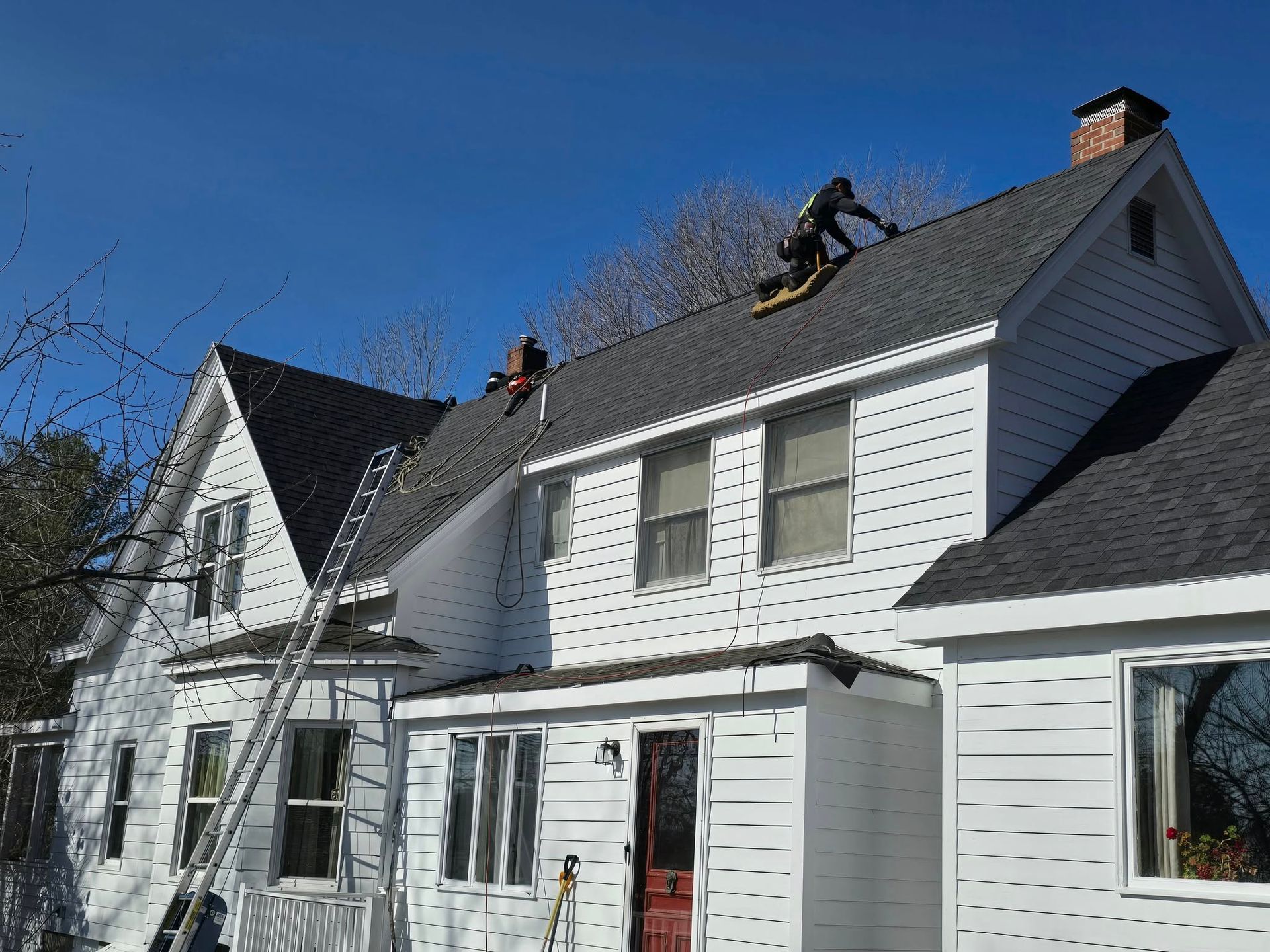Two workers on a ladder and roof of a white house with gray shingles under a clear blue sky