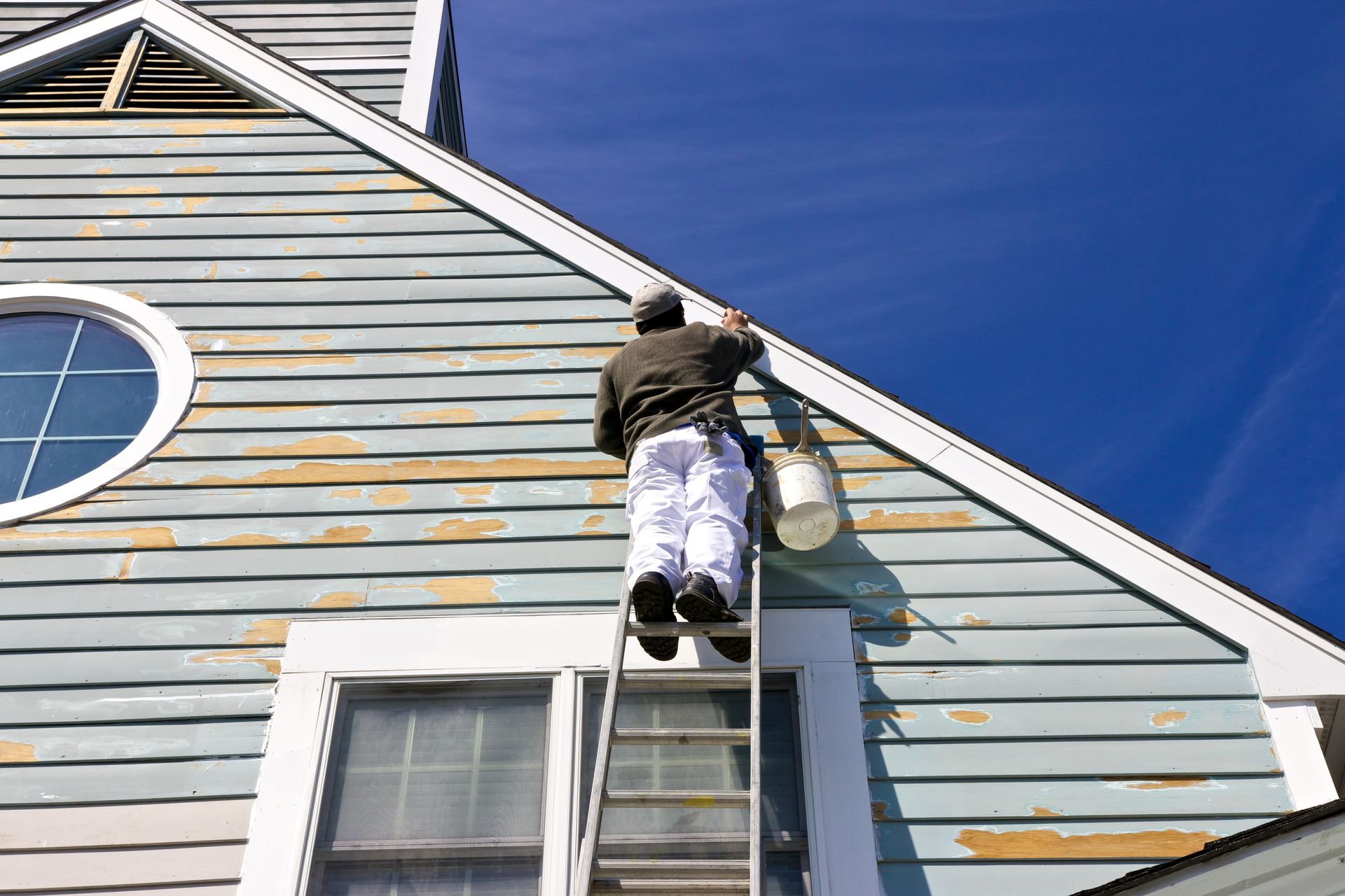 Person on a ladder painting the side of a house under a blue sky
