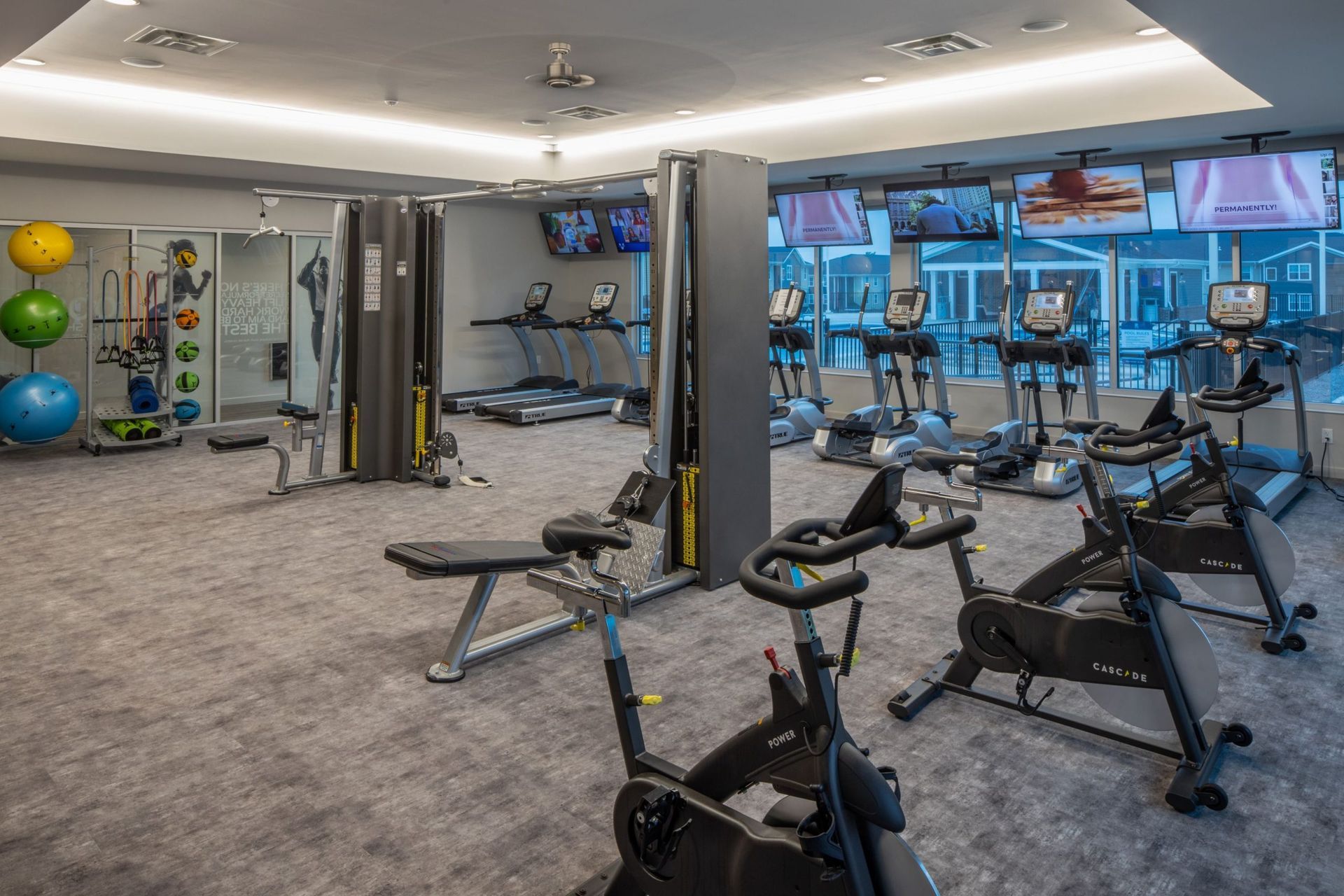Gym with cardio machines, weights, and exercise equipment. Gray carpet and bright lighting.
