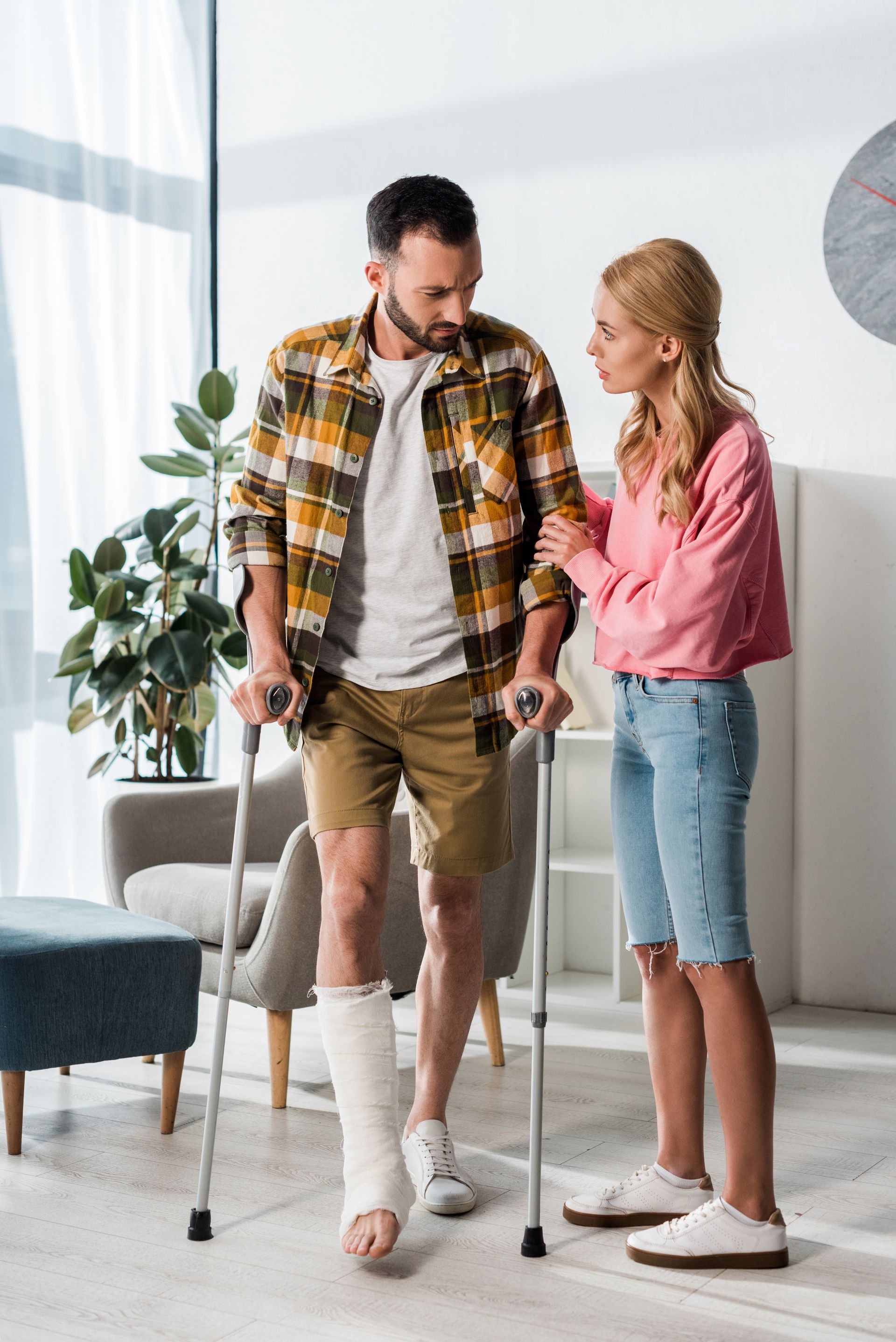 Man with a leg cast using crutches, assisted by a woman in a home setting.