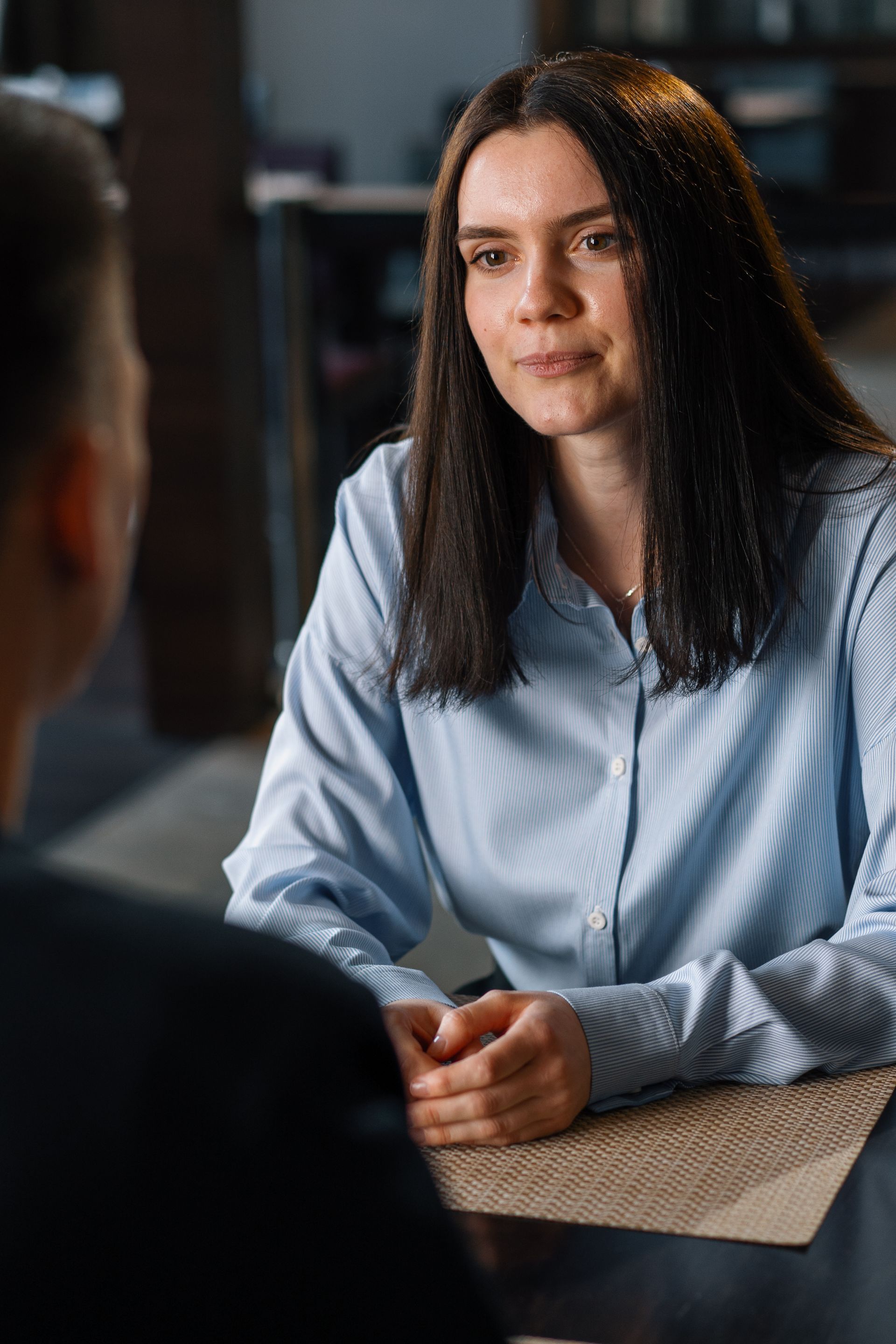 Woman in blue shirt looking intently at another person, seated at a table.