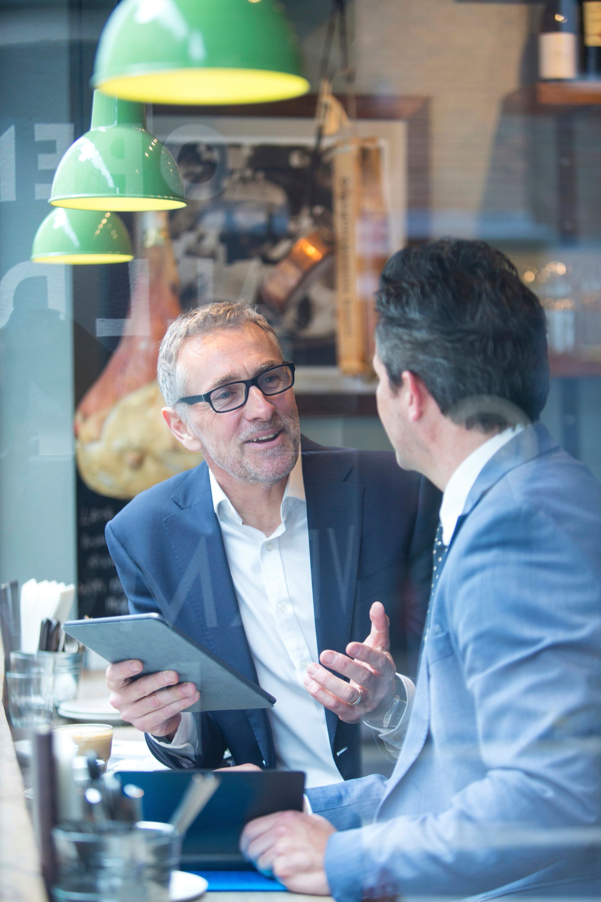 Two men in suits talking at a cafe, one holding a tablet, discussing; reflected window view.