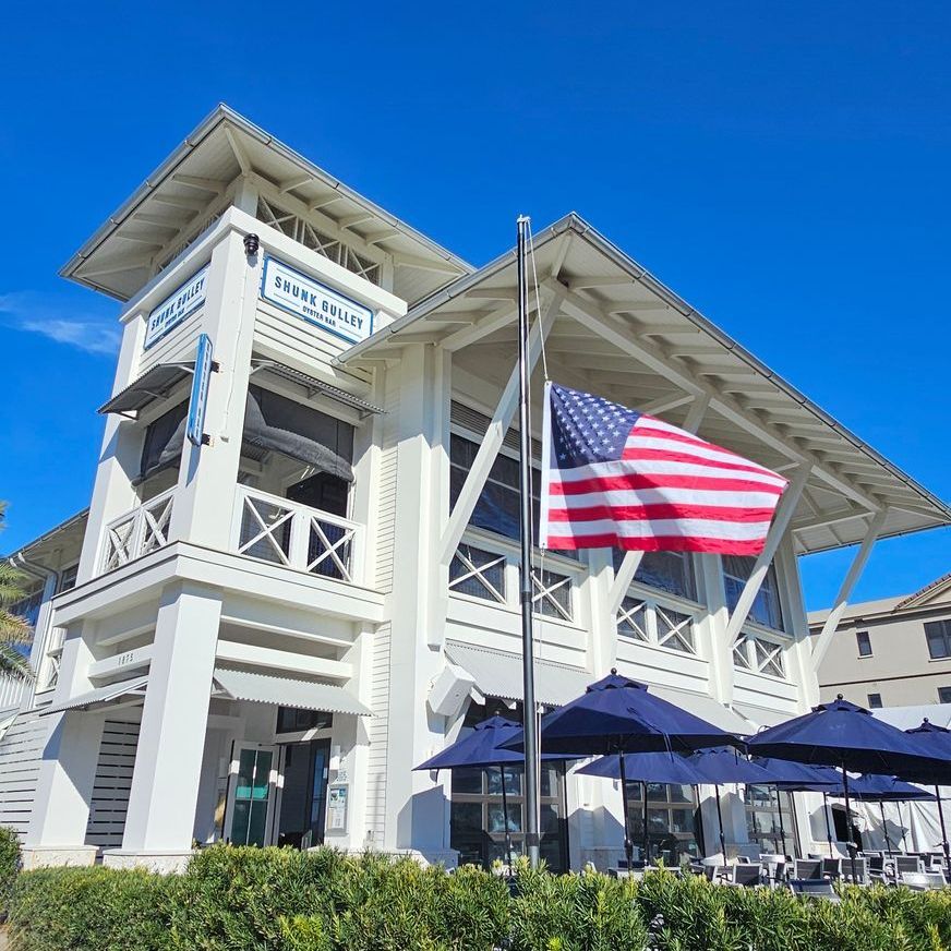 White coastal-style building with balconies, a large American flag, and blue umbrellas on a sunny day