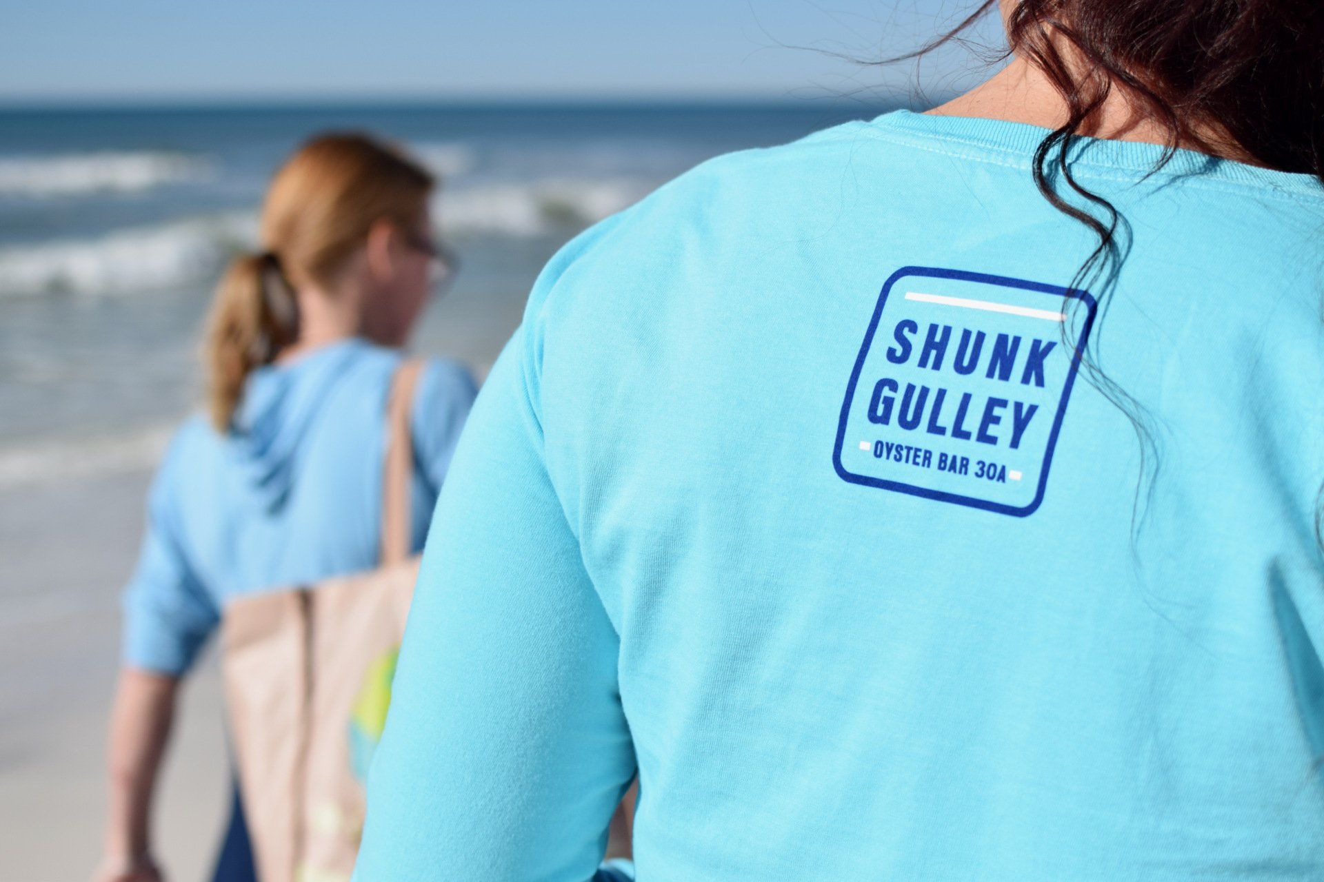 People in blue shirts walking on a beach, with a close-up of a Shunk Gulley logo on one shirt