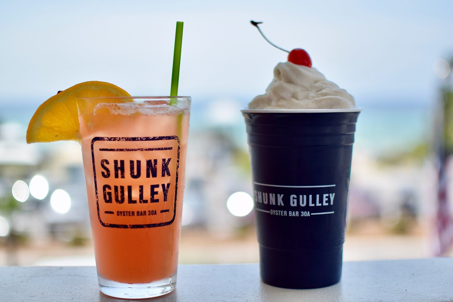 Two colorful drinks with SHUNK GULLEY logos, one orange with an umbrella and one dark with whipped cream and a cherry.