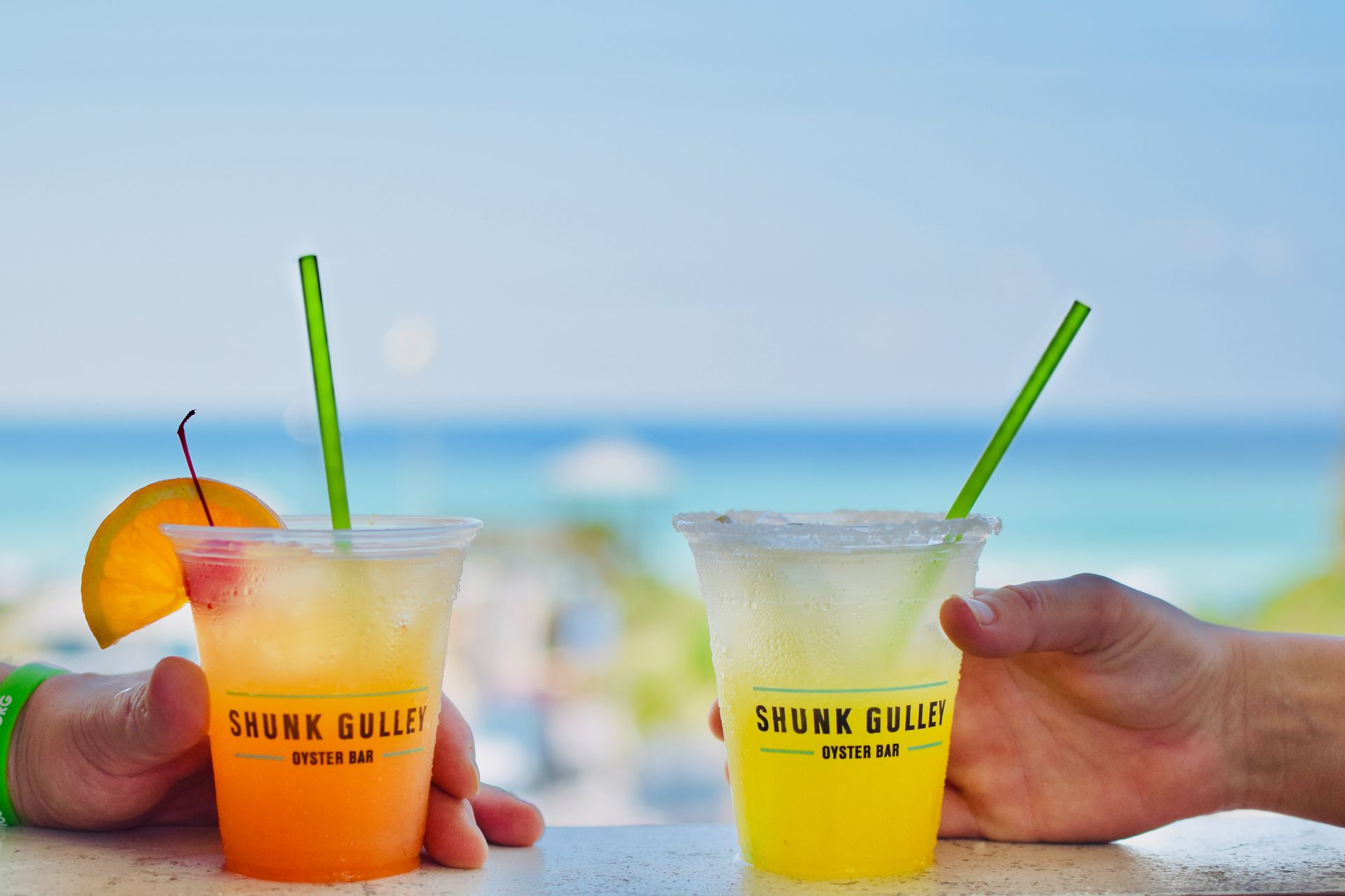 Two colorful cocktails with green straws and hands holding them on a sunny beach.