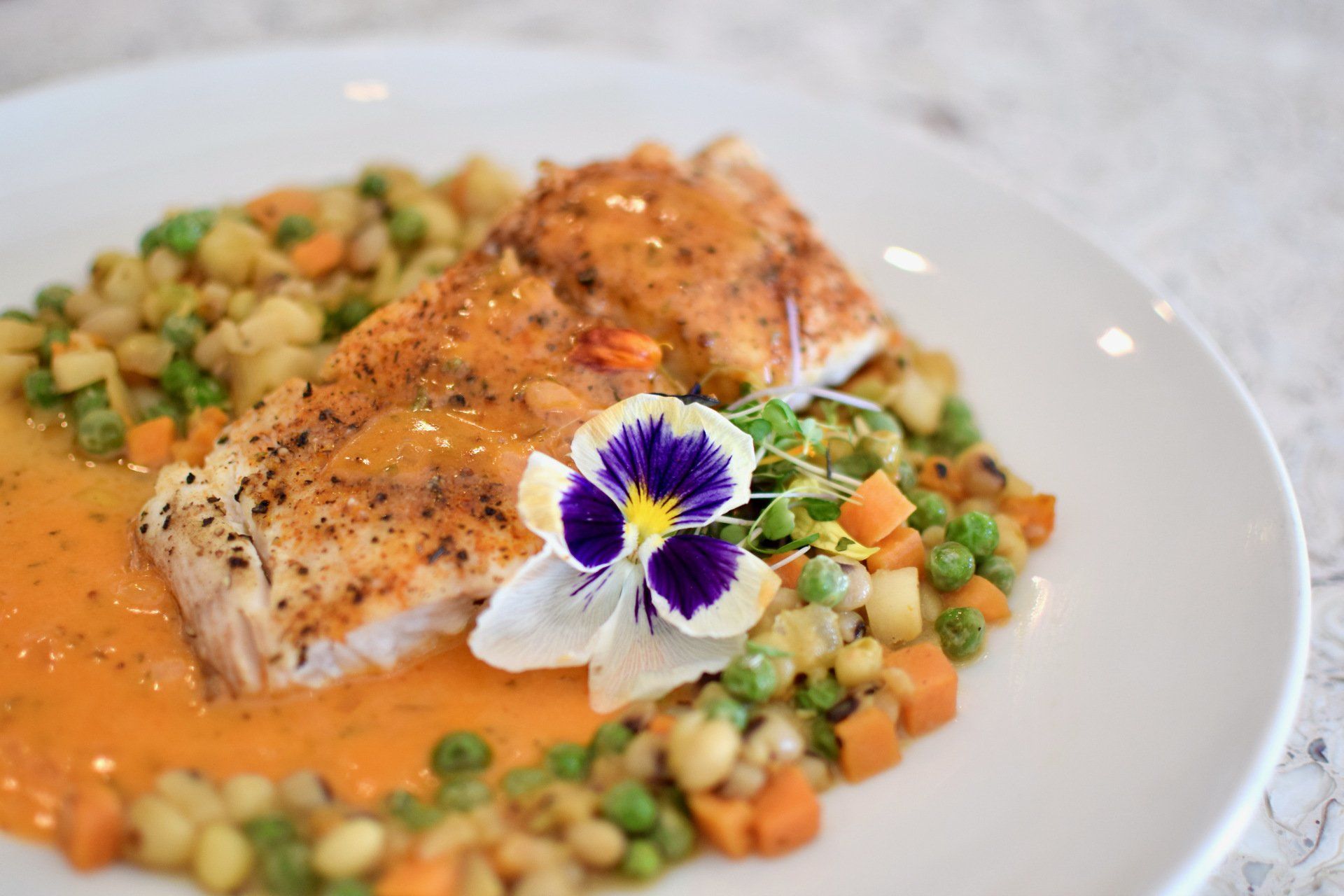 Seared salmon with peas, diced vegetables, and orange sauce, garnished with a purple edible flower