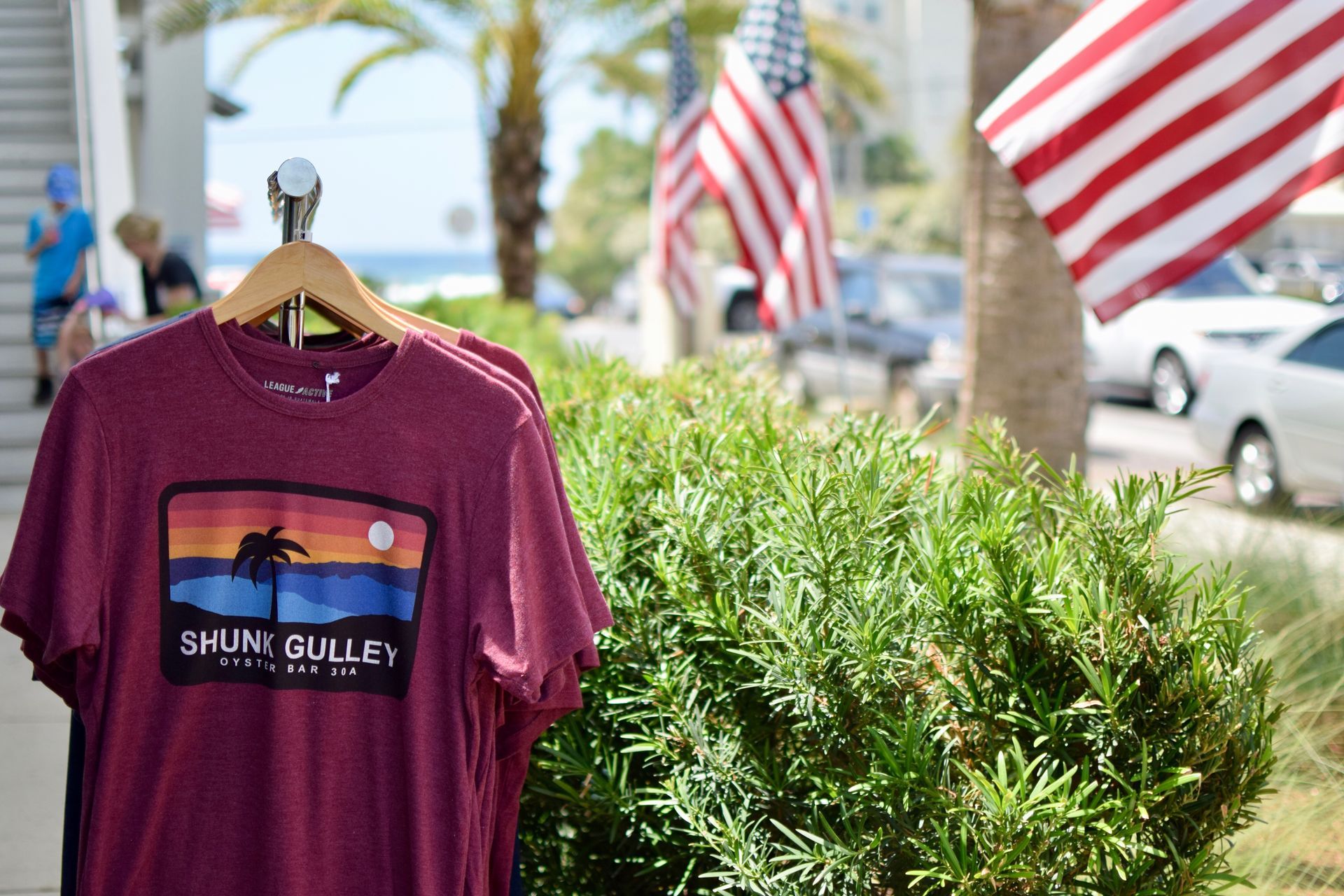 Purple T-shirt on a hanger at an outdoor market, with palm trees, flags, and cars in the background