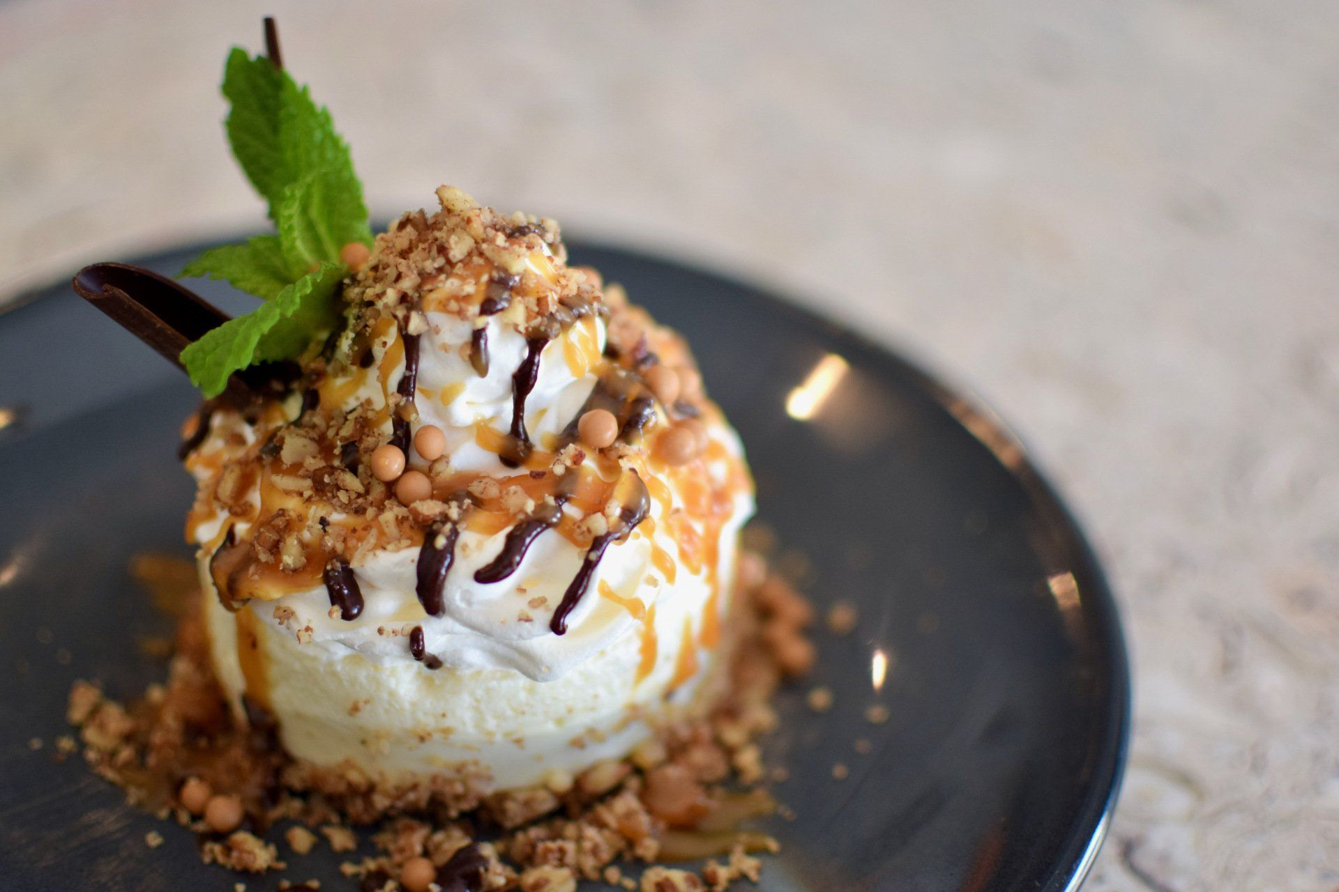 Ice cream dessert topped with nuts, chocolate drizzle, and mint on a black plate.